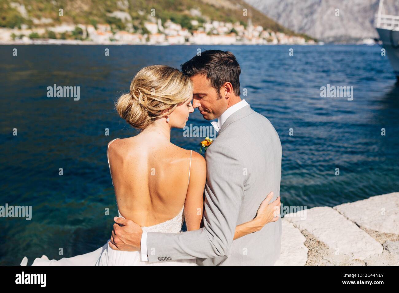 Braut und Bräutigam sitzen umarmt auf dem Pier in der Bucht von Kotor, vor ihnen ist die Altstadt, aus nächster Nähe Stockfoto