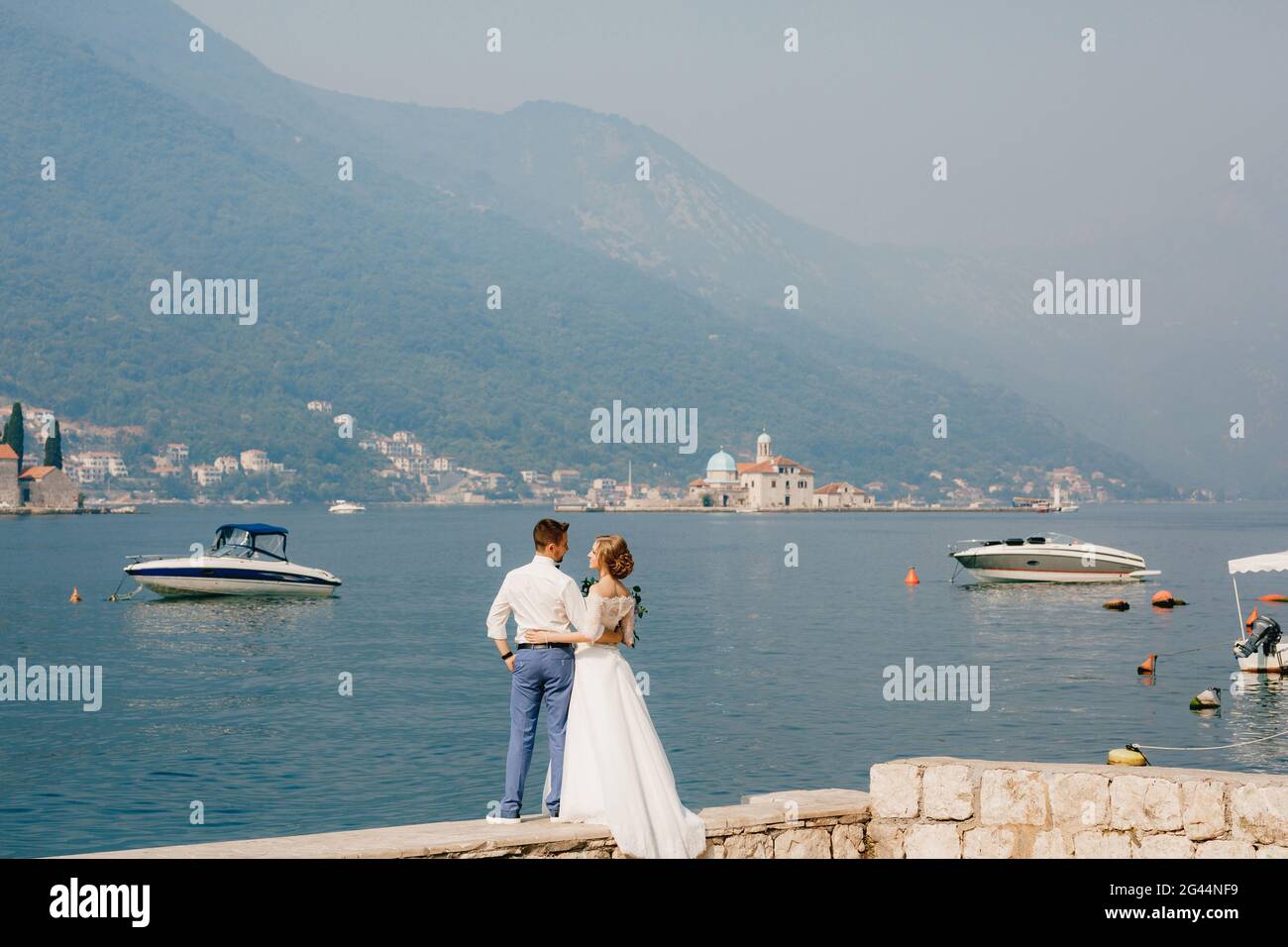 Die Braut und der Bräutigam umarmen sich auf dem Pier in der Nähe der Altstadt von Perast, Boote schwimmen in der Nähe Stockfoto