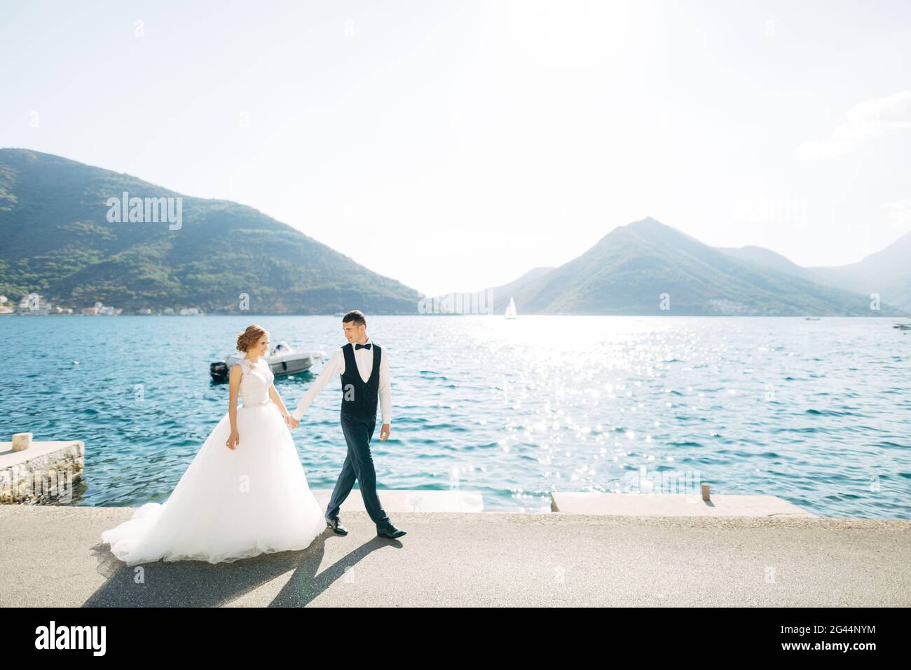 Die Braut und der Bräutigam gehen Hand in Hand auf der Straße am Meer, hinter ihnen liegen Berge und ein Boot Stockfoto