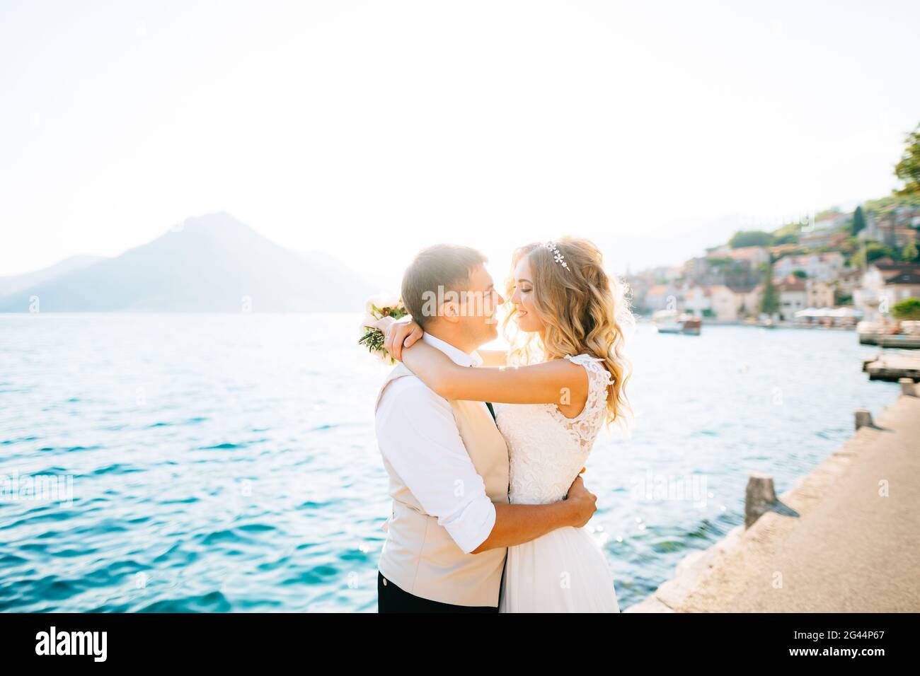 Die Braut umarmt den Bräutigam und hält einen Strauß in sich Ihre Hände auf dem Pier in der Bucht von Kotor Stockfoto