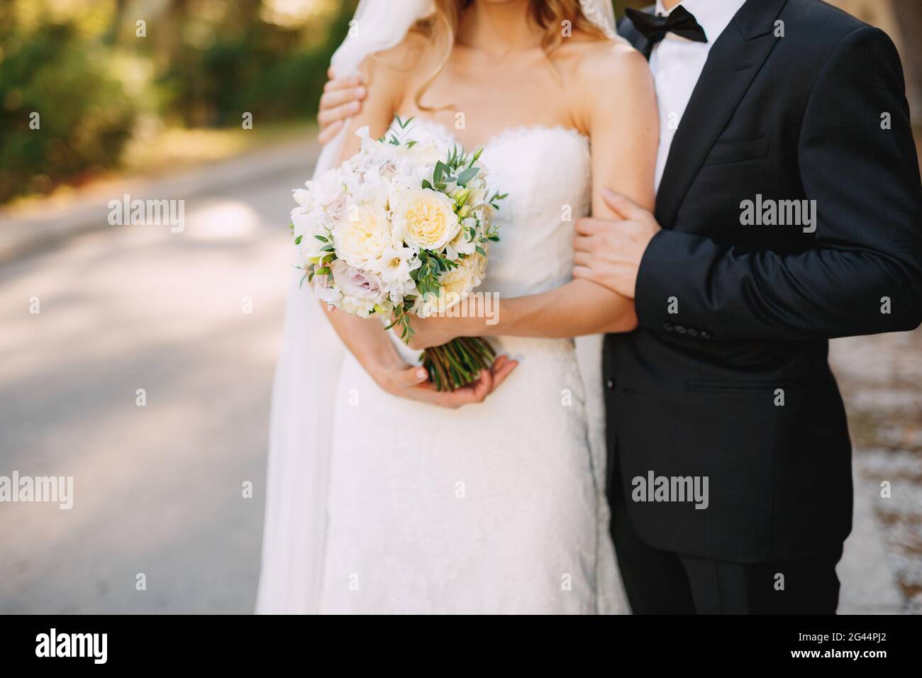 Der Bräutigam umarmt die Schultern der Braut, die einen Hochzeitsstrauß mit weißen Blumen in der Nähe hält Stockfoto
