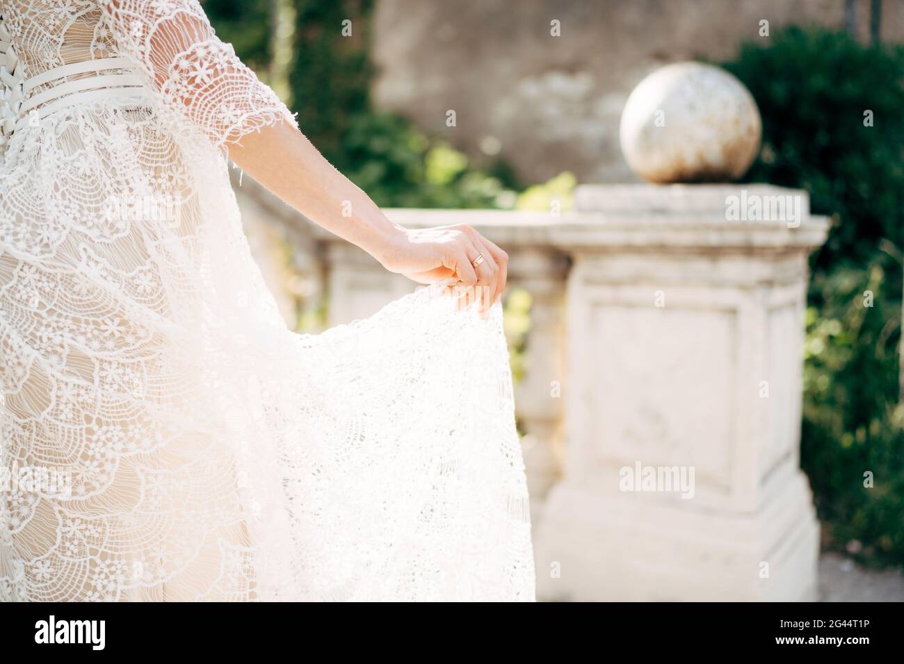 Die Braut hält mit ihren Fingern den Boden eines Weißen Spitze Hochzeitskleid auf dem Hintergrund der Säulen in Der Garten Stockfoto