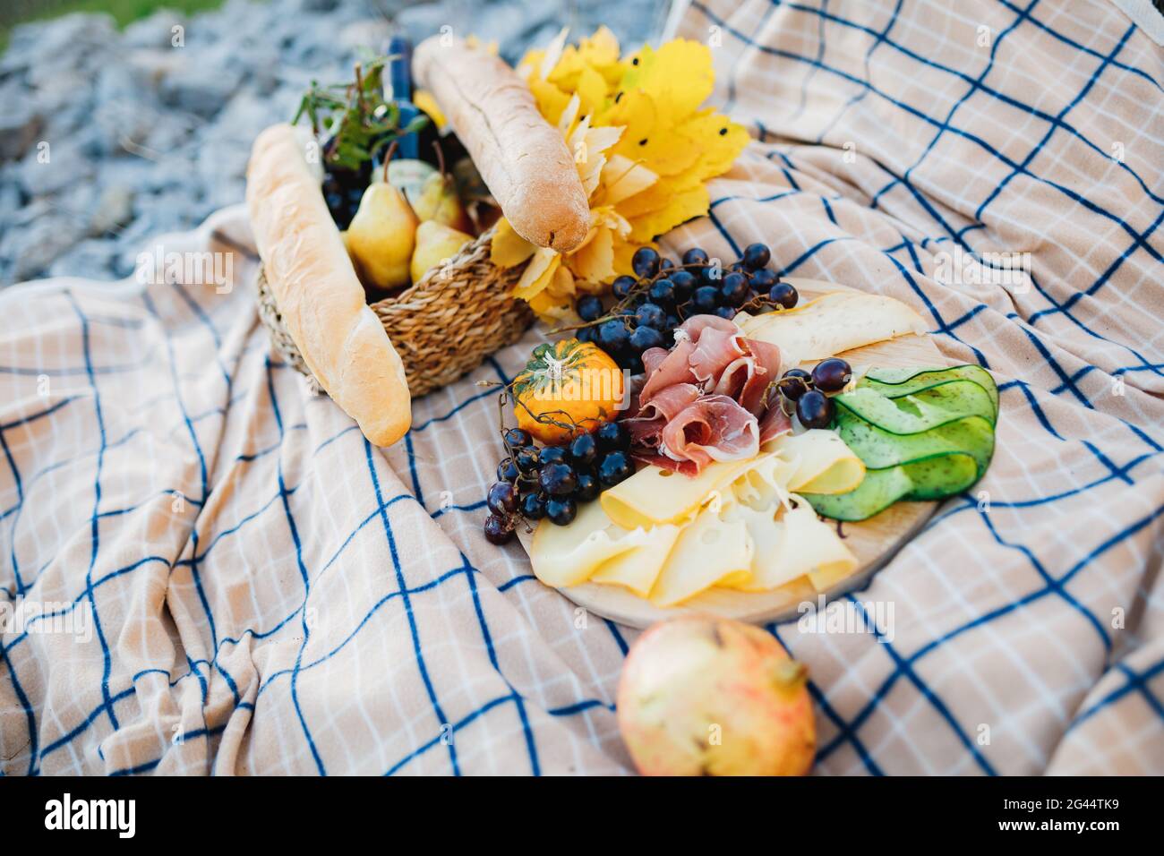 Picknick im Freien mit einem Teller mit Fleisch- und Käseschnitten und Weintrauben, einem Korb mit Baguette und einer Flasche Wein auf einer Matte. Stockfoto