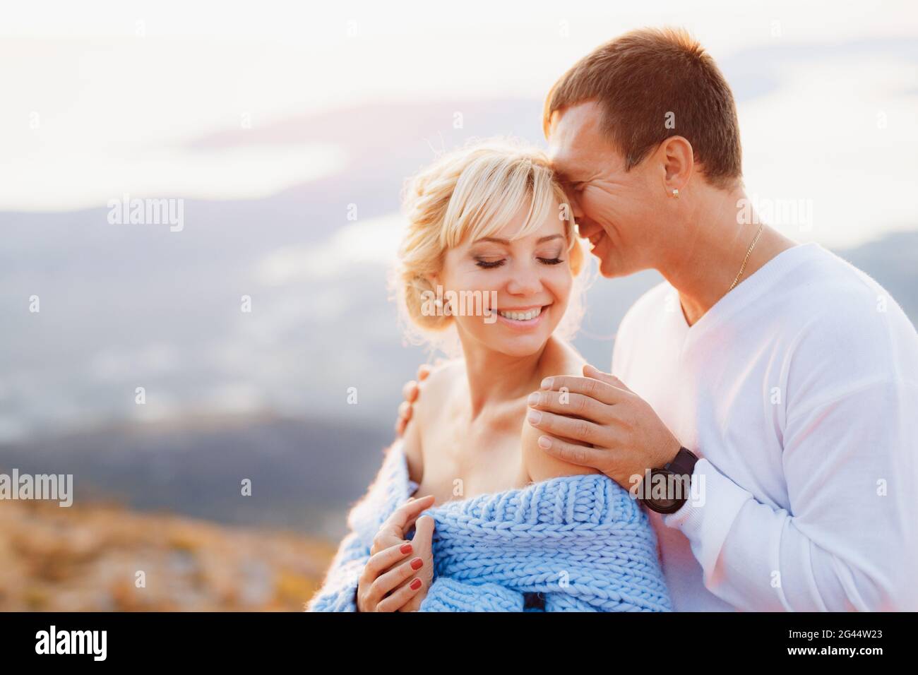 Lächelnder Mann umarmt lächelnde Frau in einem blauen Pullover an den Schultern auf dem Hintergrund der Berge. Nahaufnahme Stockfoto