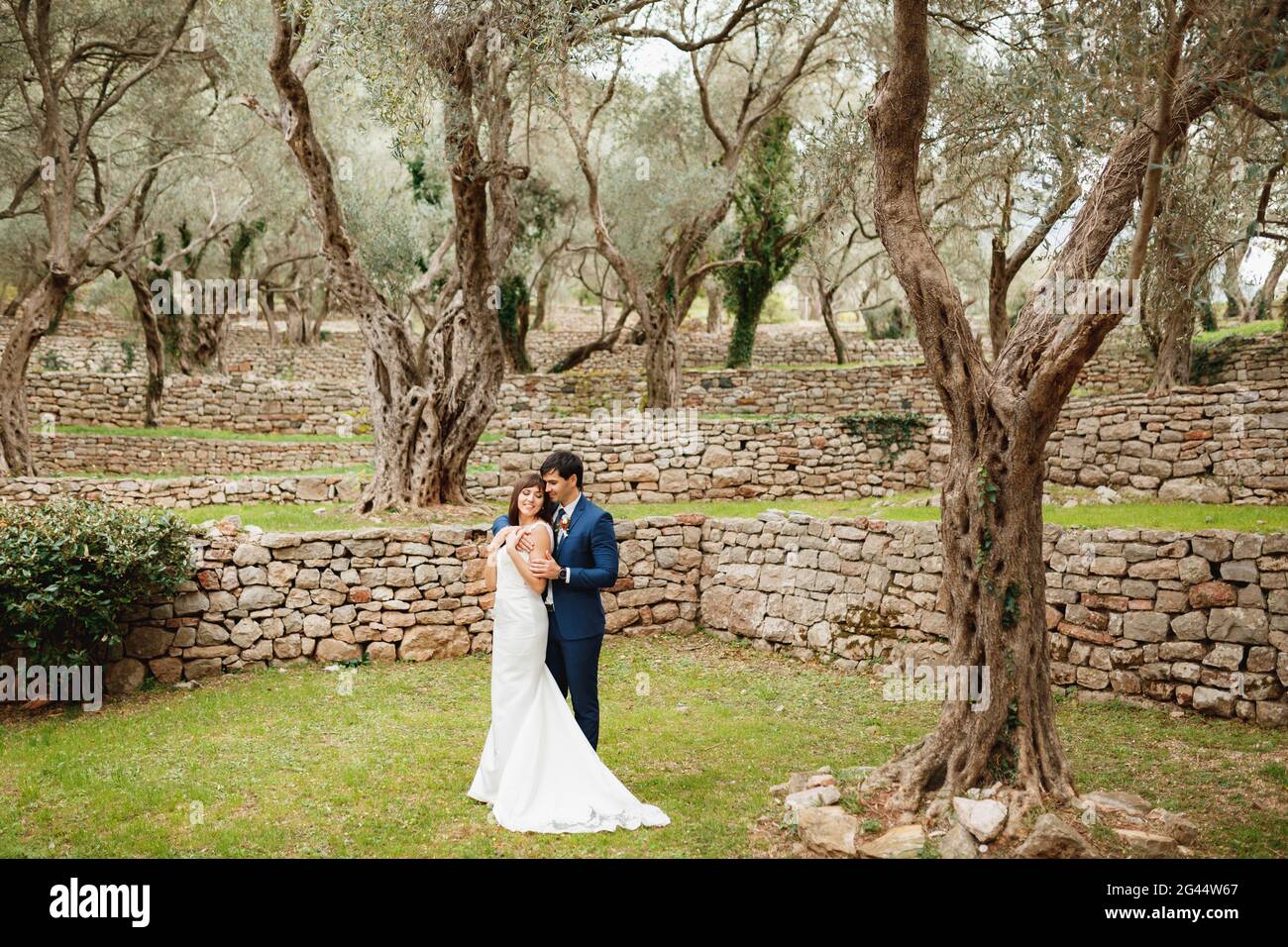 Der Bräutigam umarmt die Braut in einem weißen Hochzeitskleid vor dem Hintergrund eines wunderschönen mehrstufigen Gartens mit einem Steinzaun und einem grünen T Stockfoto