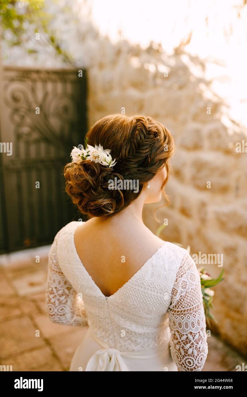 Stilvolle Braut steht zurück in einem weißen Spitzenkleid mit Eine luxuriöse Frisur und Blumen im Haar sieht an Die gegenüberliegende Wand Stockfoto