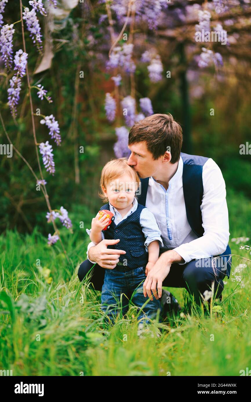 Vater küsst sein Baby Junge auf der Oberseite der Kopf mit einer im Hintergrund blühenden Wysterie Stockfoto