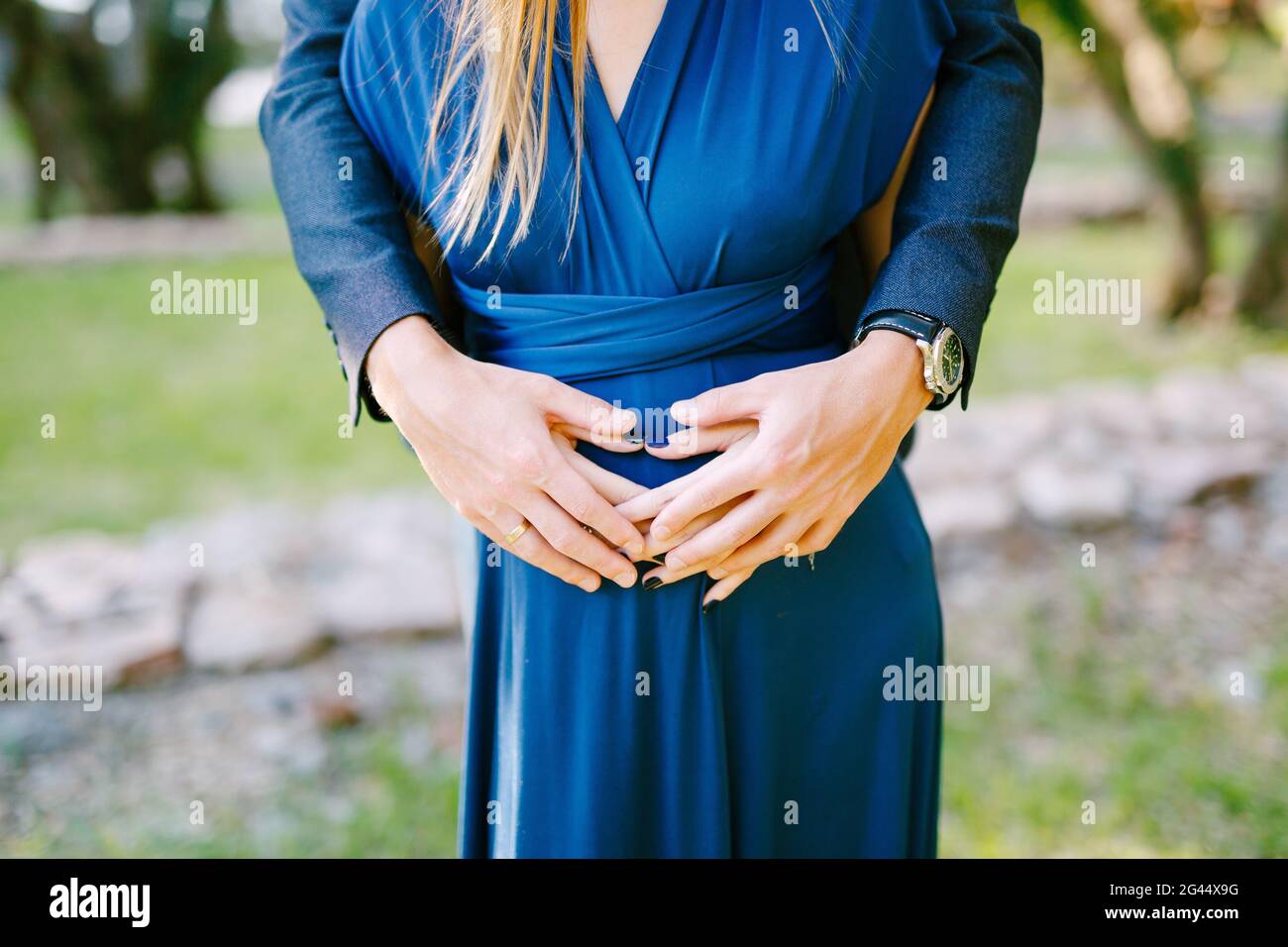 Die Hände des Mannes umarmen sich hinter dem schwanger Bauch der Frau In einem blauen Kleid Stockfoto