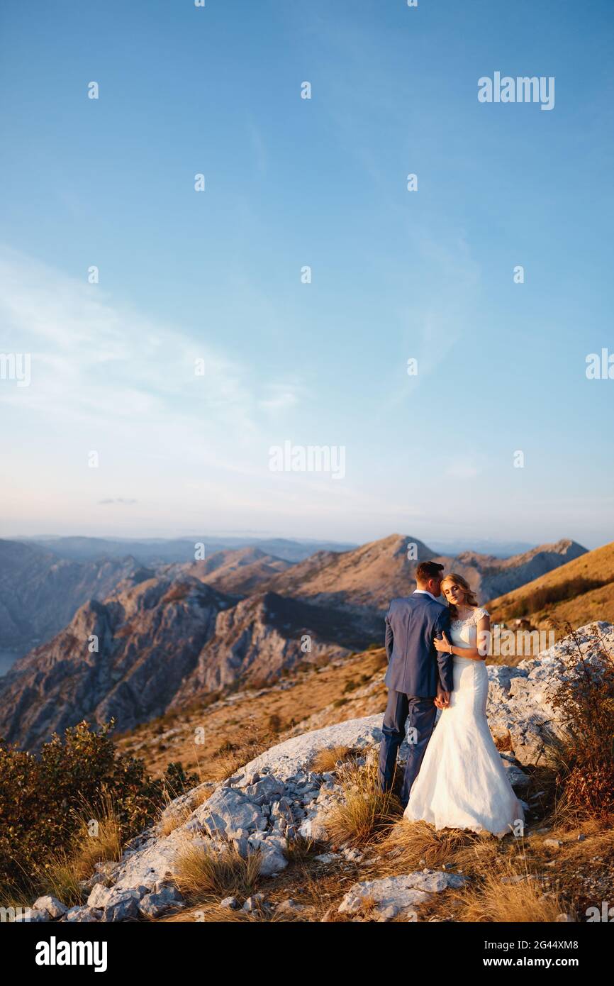 Die Braut umarmt den Bräutigam und hält seine Hand auf dem Gipfel des Mount Lovcen mit Blick auf die Bucht von Kotor, lächelt und umarmt zärtlich Stockfoto