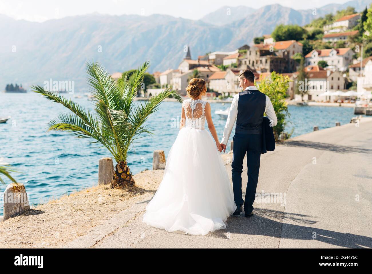 Braut und Bräutigam gehen Hand in Hand auf der Straße am Meer in der Nähe der Altstadt von Perast, Rückansicht Stockfoto