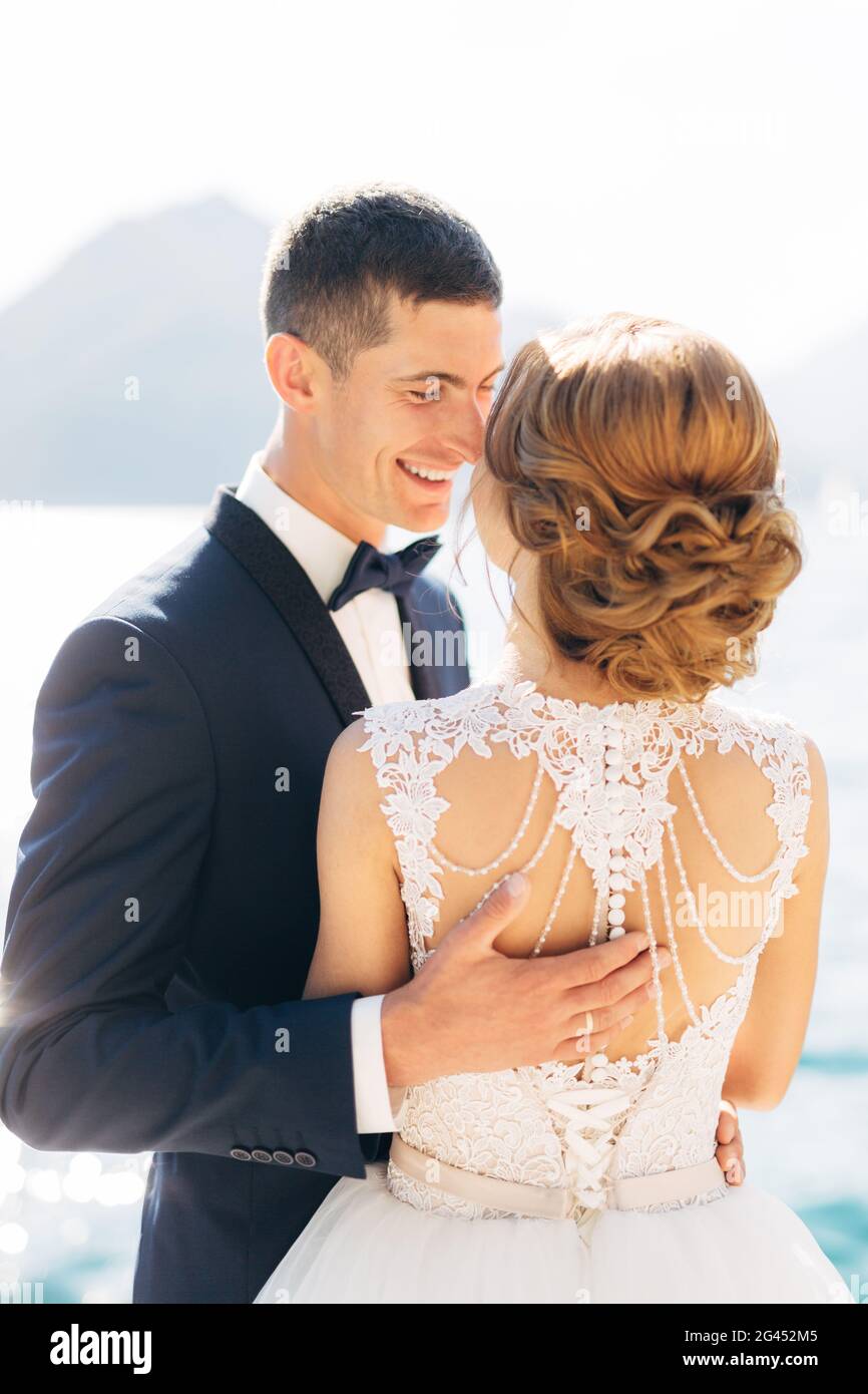 Die Braut und der Bräutigam stehen, umarmen und lächeln auf dem Pier in der Nähe des Meeres, aus der Nähe Stockfoto