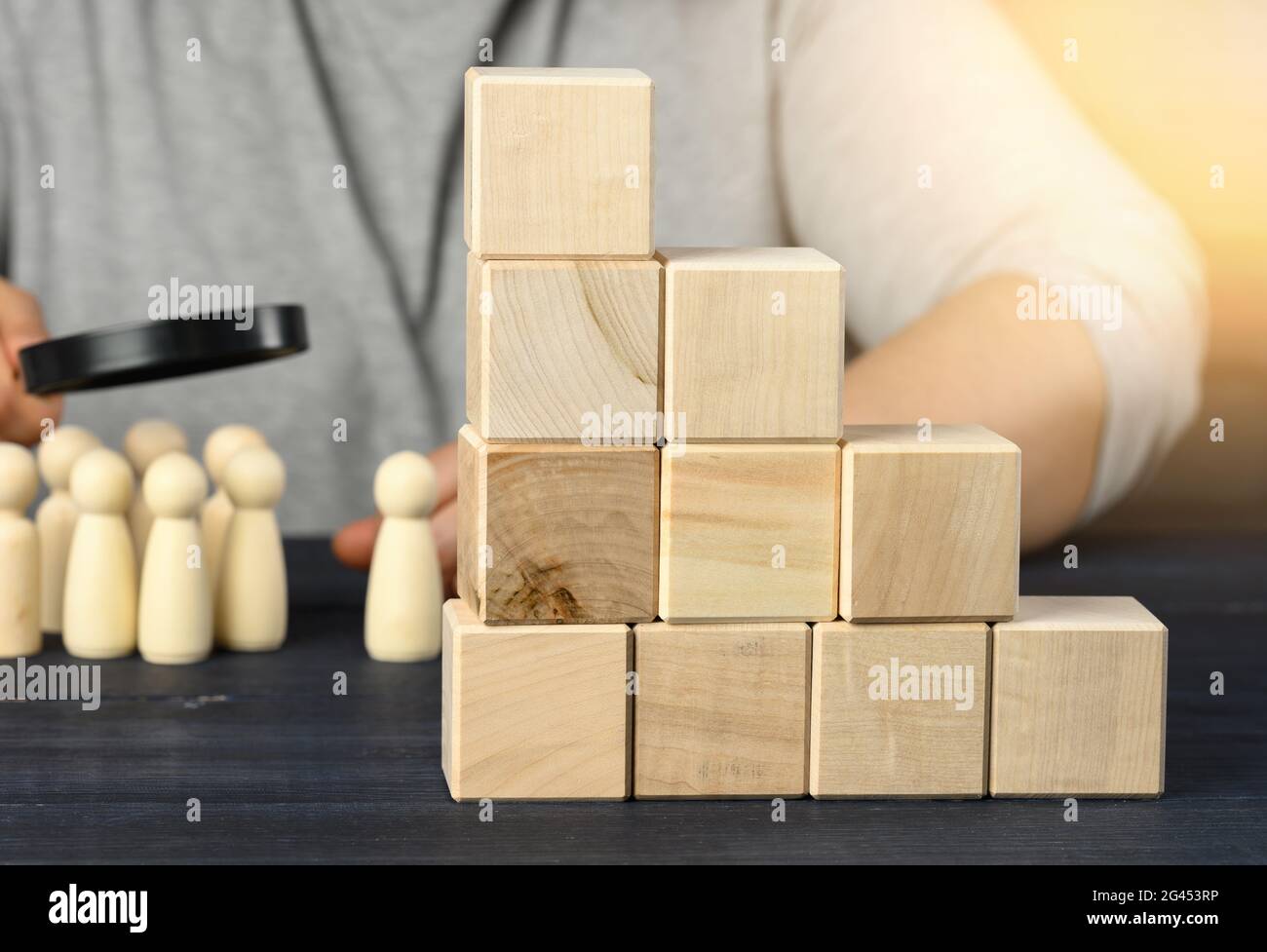 Leiter aus Holzwürfeln auf einem Stahl, hinter einem Mann unter einer Lupe untersucht Holzfiguren. Rekrutierungskonzept Stockfoto