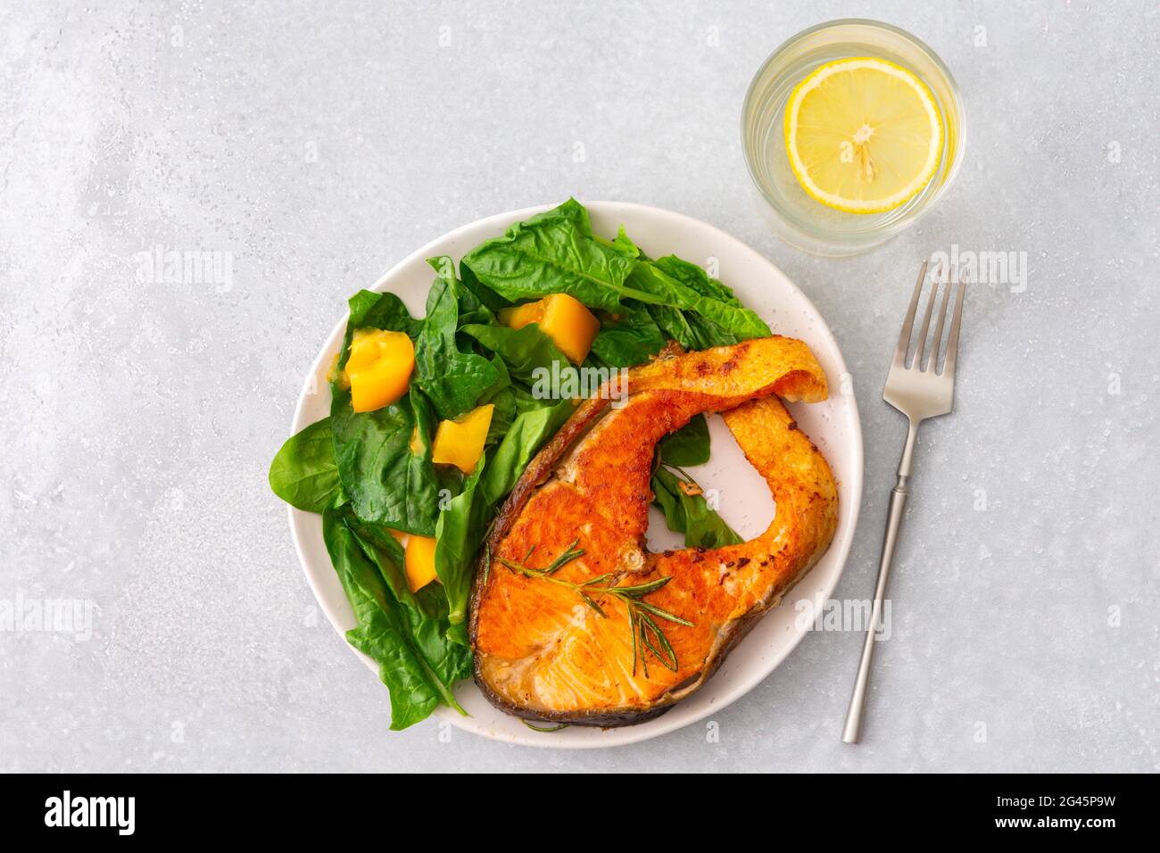 Gebratenes Lachssteak mit Spinat und gelben Tomaten auf einem Teller auf grauem Grund, ein Glas Wasser mit Zitrone, ein herzhaftes Mittagessen mit gesundem rotem Fisch Stockfoto