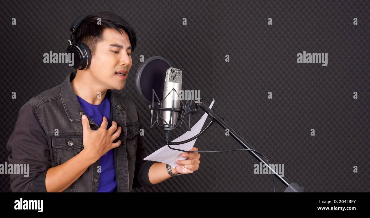 Ein junger asiatischer Mann mit Kopfhörern singt, während er vor schwarzen Schallschutzwänden die Texte in der Hand hält. Musiker, die Musik im professionellen Stockfoto