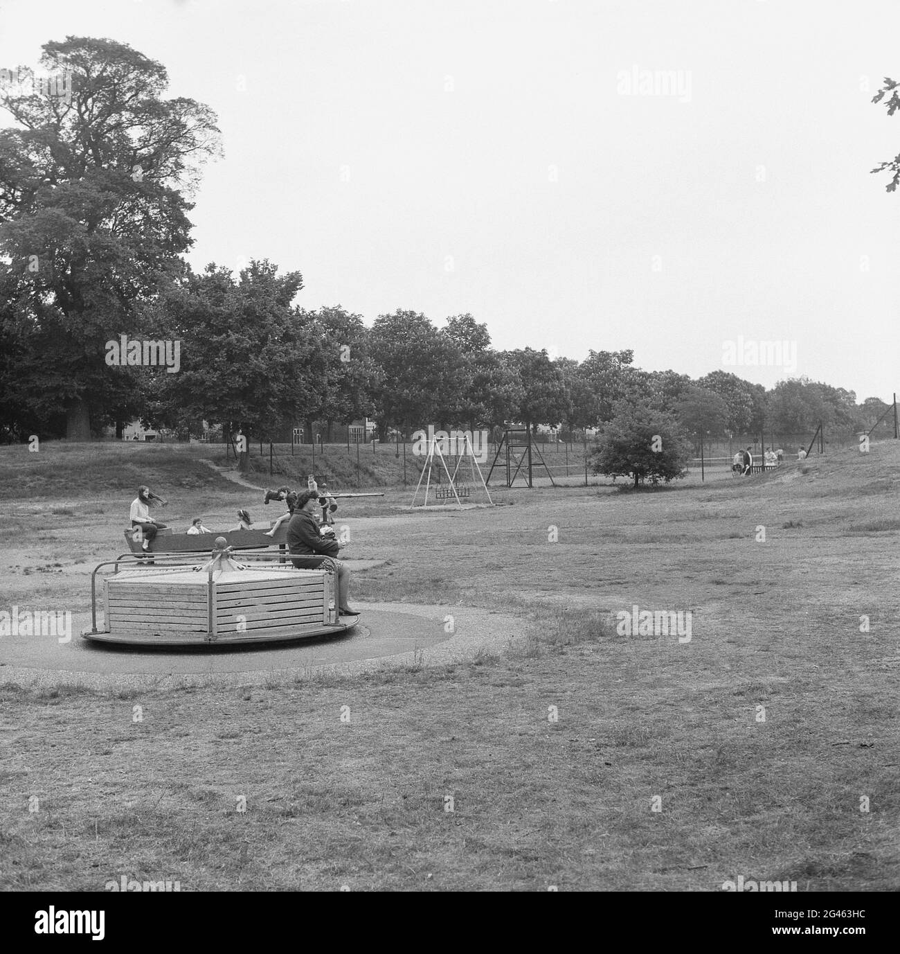 1969, historische, Kinder auf einem Spielplatz in einem öffentlichen Park, zeigt die Spielgeräte des Tages; ein Karussell oder ein Karussell, ein Metall-Spinnrad, angetrieben von den spielenden Kindern und ein Metall-Schaukelpferd, alle später verboten, als zu gefährlich angesehen, England, Großbritannien. Stockfoto