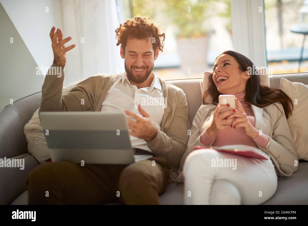 Schönes junges Paar, das auf dem Sofa lachend während der sozialen Isolation von zu Hause aus arbeitet, weil das Coronavirus gesperrt wurde und sich gut amüsiert hat Stockfoto