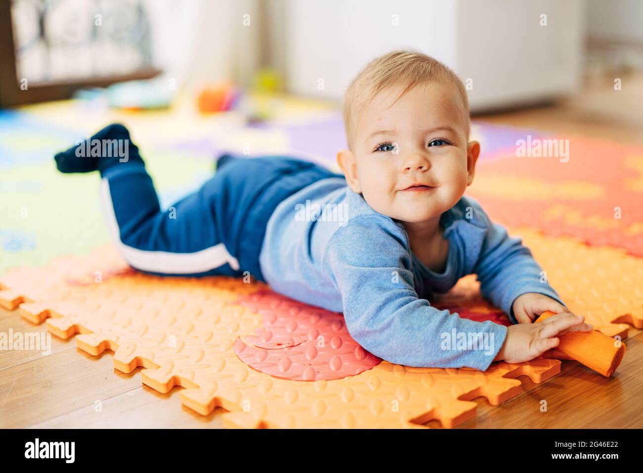 Lächelnder kleiner Junge in einem blauen Anzug mit einer Karotte in den Händen liegt mit seinem Bauch auf einem farbigen Teppich auf dem Boden gegen den Stockfoto