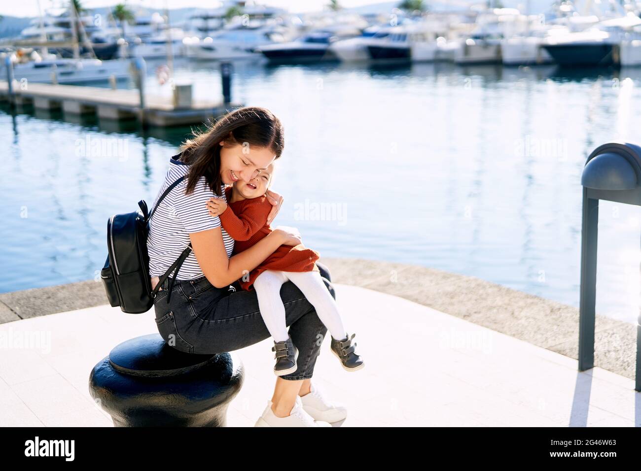 Junge Frau umarmt ihre Tochter fest, während sie darauf sitzt Ein Pier in einer Marina Stockfoto
