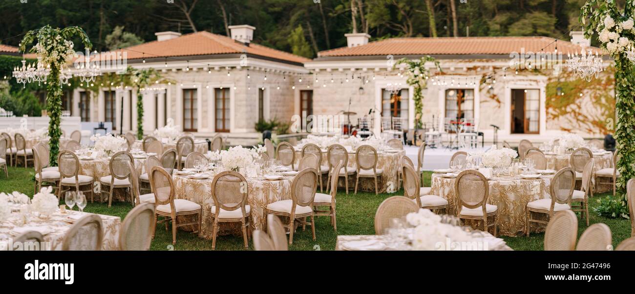 Hochzeitsessen Tischempfang. Elegante Tische für Gäste mit cremefarbenen Tischdecken mit Mustern, auf grünem Rasen, mit Girlanden und Stockfoto