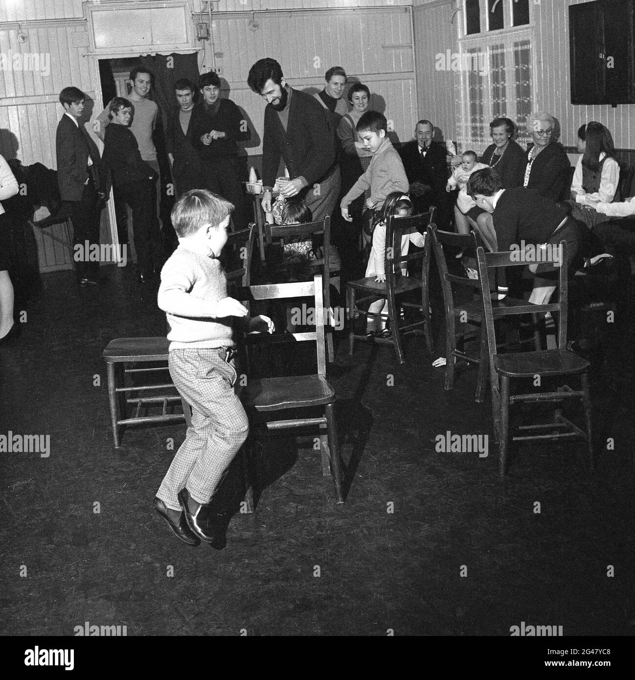 In Essex, England, Großbritannien, sehen Erwachsene in einer Dorfhalle, wie kleine Kinder auf einer Familienfeier auf Holzstühlen ein Spiel spielen. Stockfoto