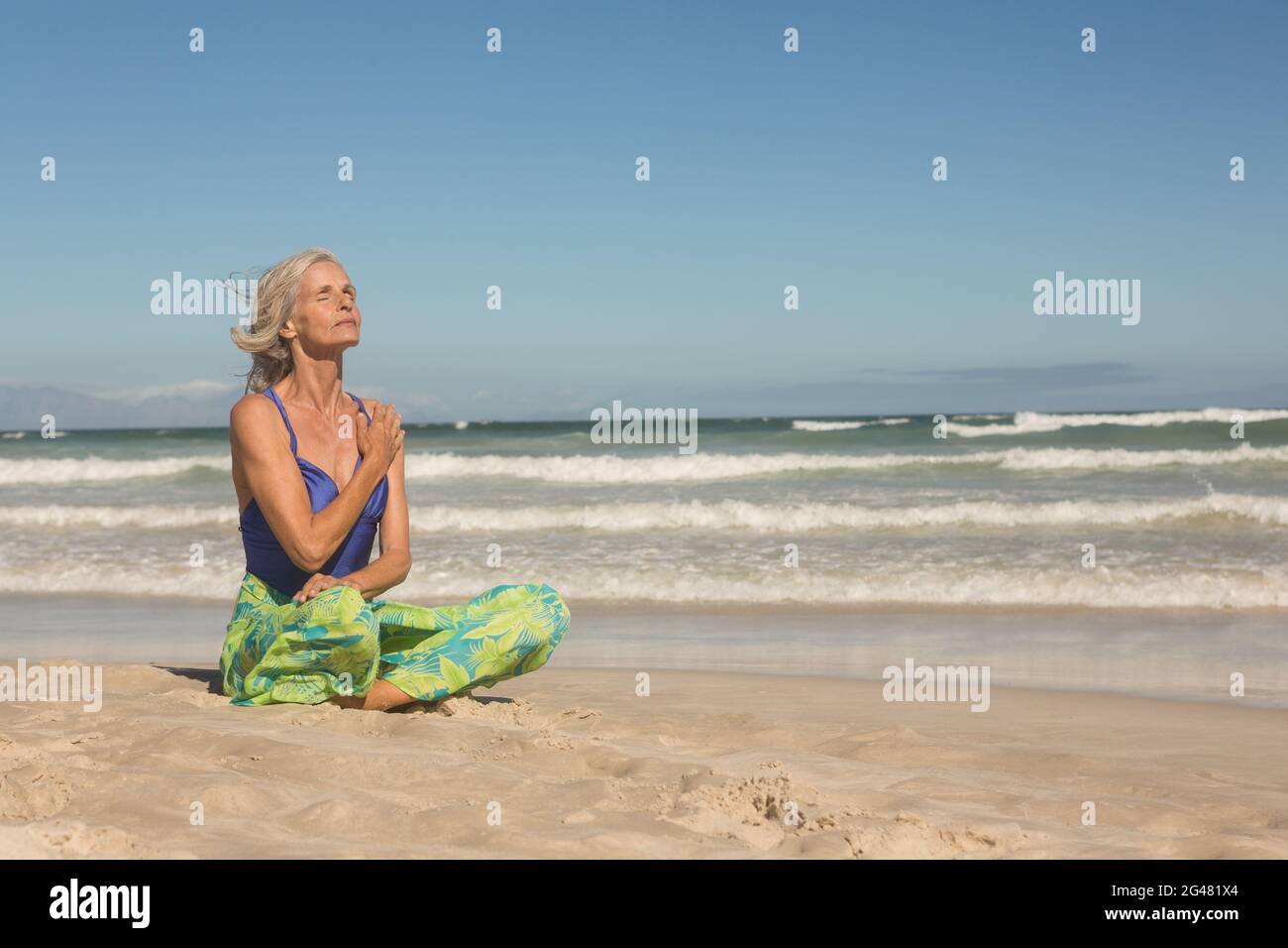 Ältere Frau meditiert, während sie am Ufer gegen den klaren Himmel sitzt Stockfoto