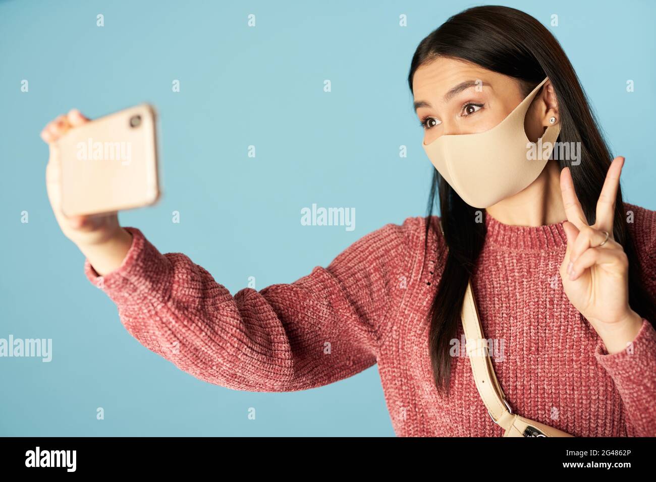 Lady in Maske macht Foto auf dem Smartphone und zeigt Siegeszeichen Stockfoto