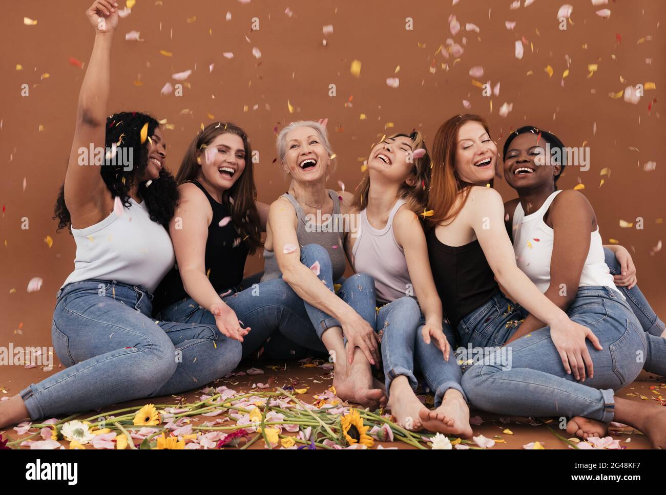 Gruppe von glücklichen Frauen auf dem braunen Hintergrund sitzen, während Blütenblätter von Blumen fallen. Lachende Weibchen verschiedener Rassen und Altersgruppen unter Blütenblättern aus Stockfoto