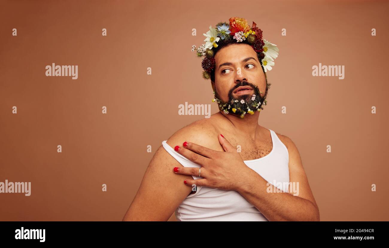 Geschlecht Queer trägt Tank Top und Blumen. Nicht binäre Person mit Make-up, die auf braunem Hintergrund wegschaut. Stockfoto