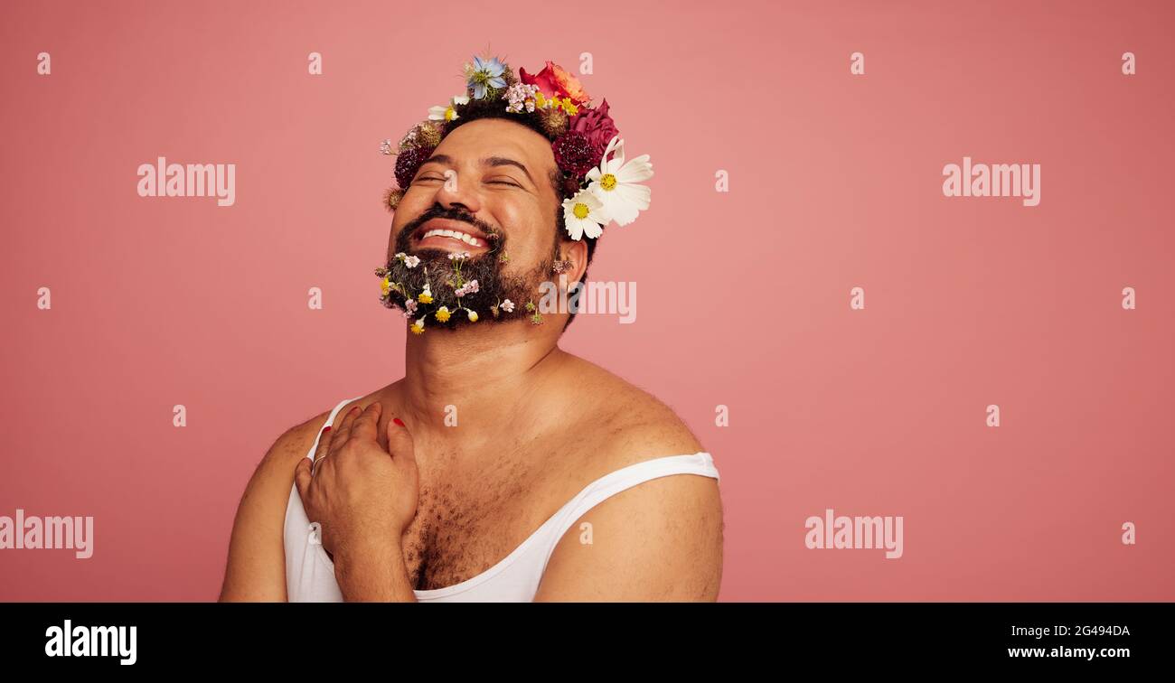 Genderqueer lächelnd auf rosa Hintergrund. Schwuler Mann trägt Make-up und Blumen auf Kopf und Bart mit Kopieplatz. Stockfoto