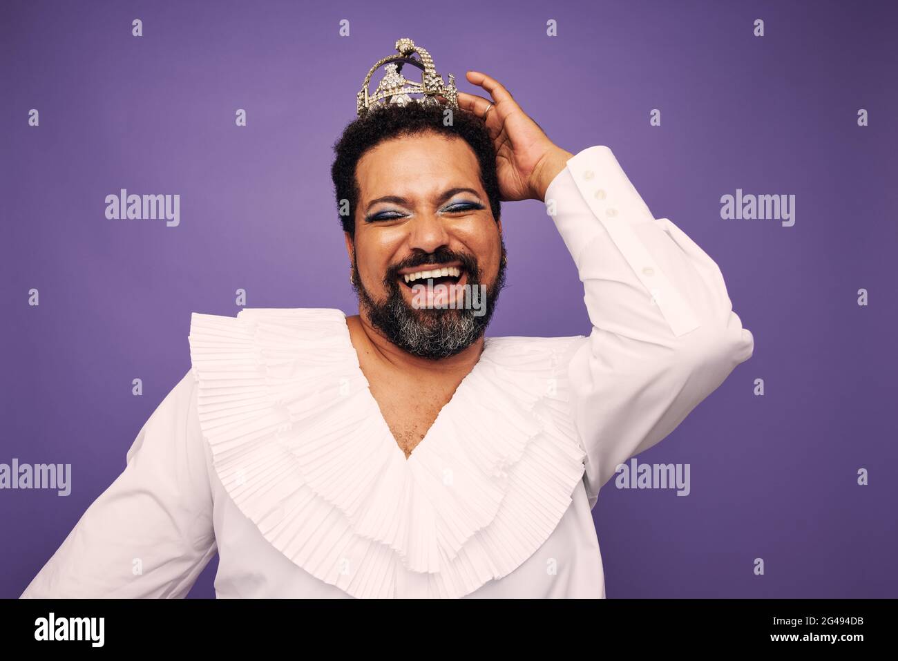 Porträt einer Drag Queen lächelt auf violettem Hintergrund. Bärtiger Mann mit Make-up und Krone auf dem Kopf. Stockfoto