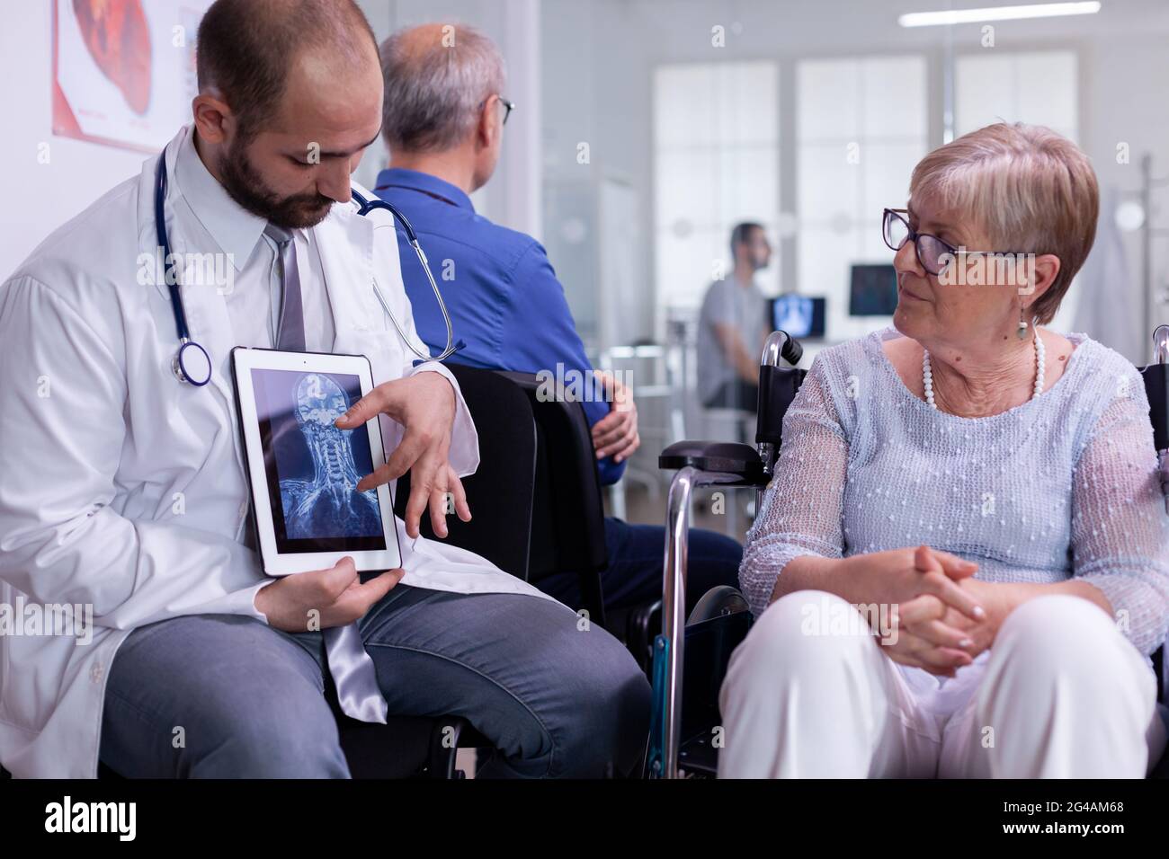 Vorderansicht eines Arztes, der mit einer behinderten älteren Frau über eine Revovery-Behandlung mit Hilfe einer Tablette sprach. Facharzt sitzt in wartenden Krankenhaus arrea Stockfoto