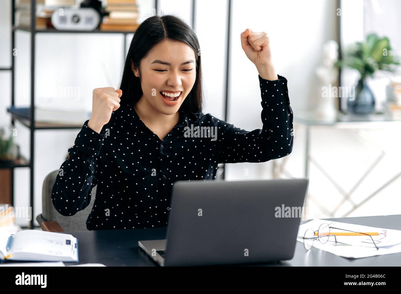 Glücklich positiv erfolgreiche asiatische junge Frau, stilvoll gekleidet, Büroangestellte, Freiberufler oder Student sitzt am Schreibtisch, gestikulierende mit ihren Händen jubelnd in Sieg, Erfolg, gutes Geschäft, großen Gewinn Stockfoto