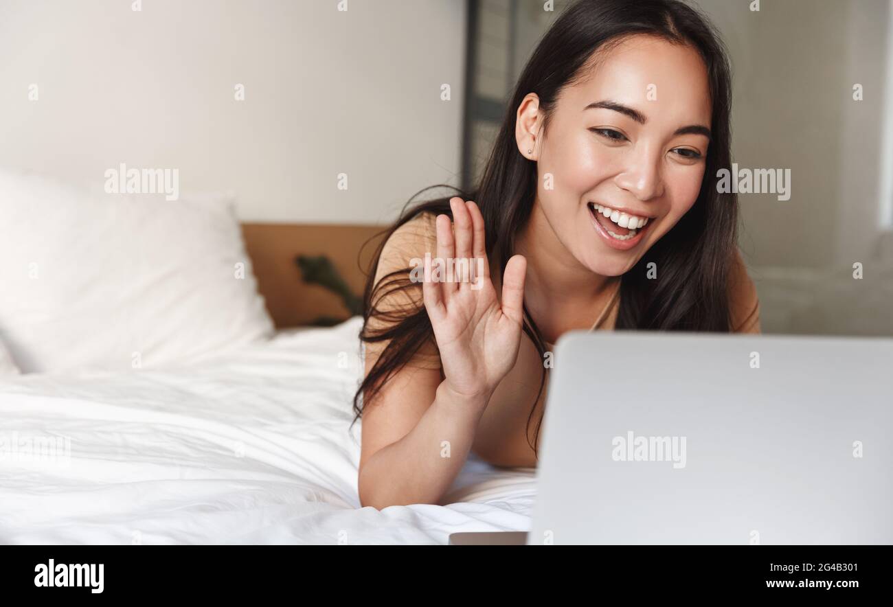 Asiatisches Mädchen liegt im Bett und Video-Chat auf dem Laptop, winkt an der Computer-Kamera, um Hallo zu sagen, grüßt Menschen Online-Konferenz, kommunizieren aus der Ferne Stockfoto