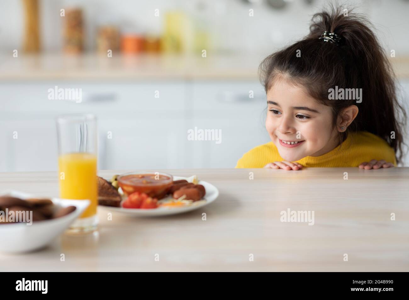 Niedliches lustiges kleines Mädchen, das unter dem Tisch guckt und auf das Essen schaut Stockfoto