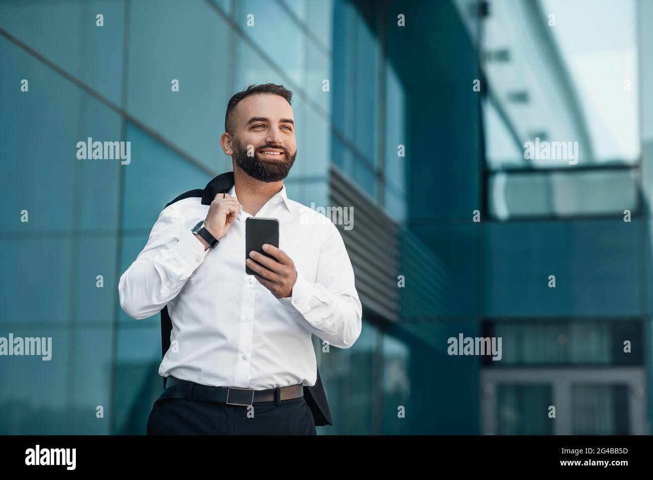 Wohlhabender, reifer Geschäftsmann, der sein Smartphone überprüft, während er in der Nähe des Bürogebäudes geht und beiseite schaut, um Platz zu kopieren Stockfoto