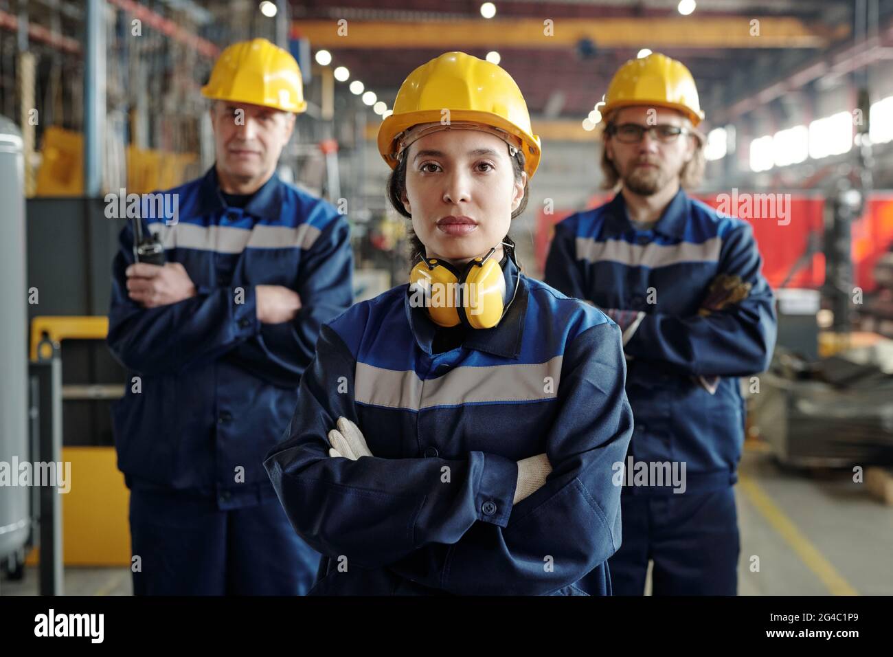 Porträt einer ernsthaften jungen Frau mit Hardhut und geräuschreduzierenden Kopfhörern, die mit männlichen Kollegen im Werk zusammenarbeitet Stockfoto
