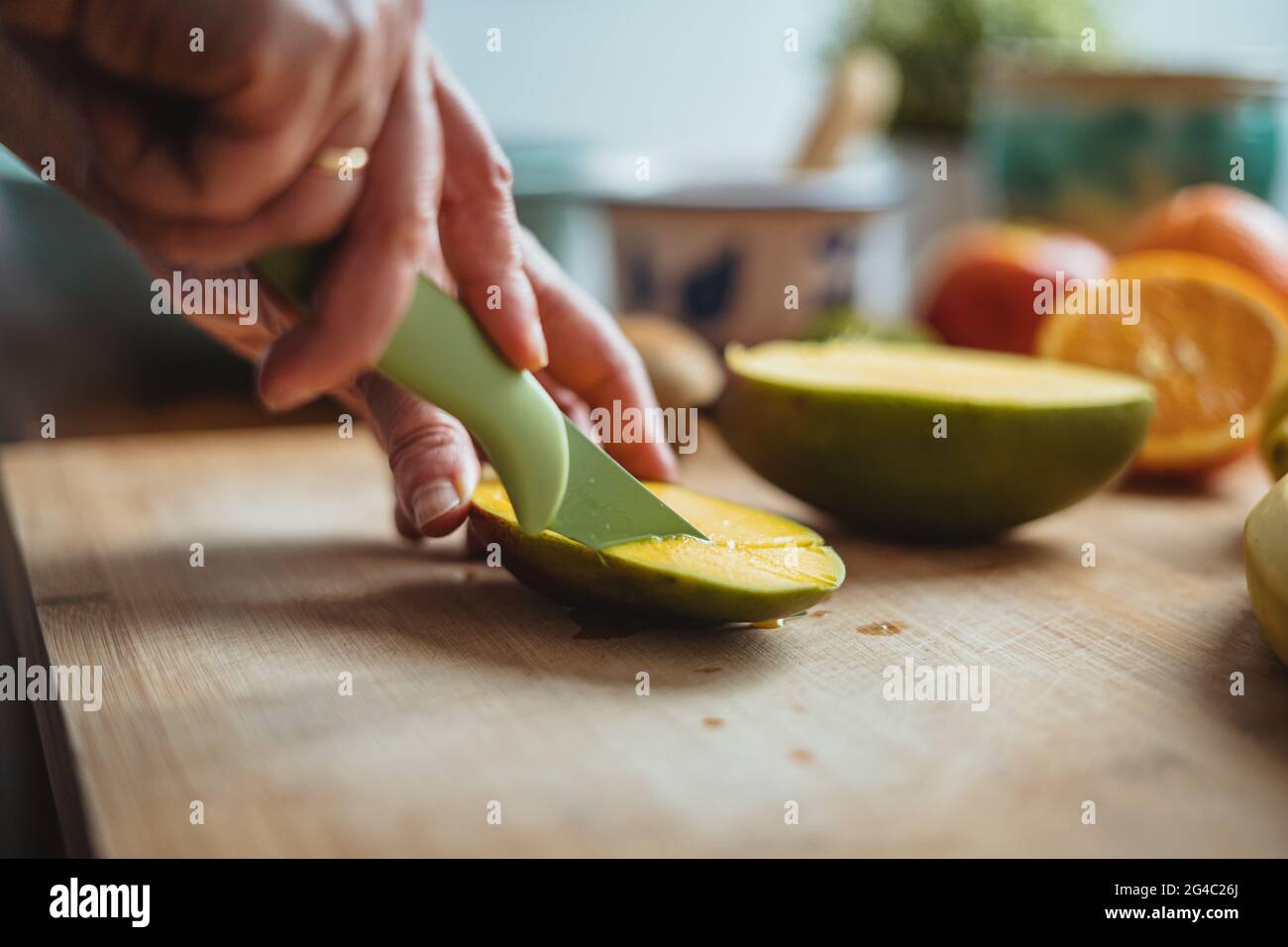 Eine Frau schneidet eine Mango mit einem grünen Messer auf einem Holztisch, der von anderen Früchten umgeben ist. Stockfoto