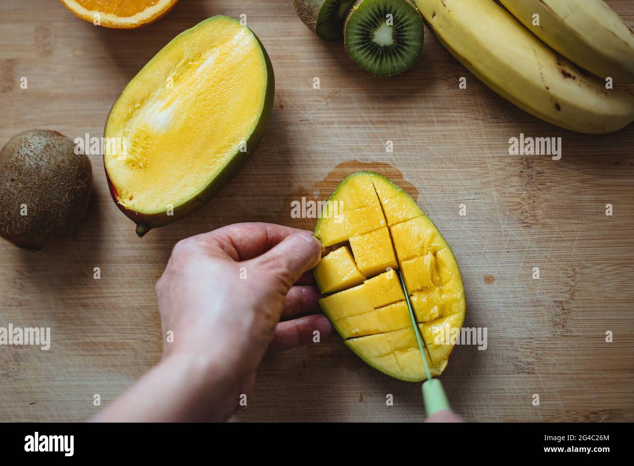Eine Frau schneidet mit einem grünen Messer eine Mango auf einem Holztisch, der von anderen Früchten umgeben ist (Draufsicht). Stockfoto