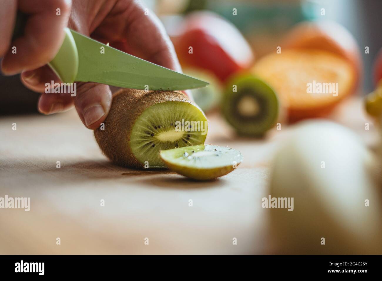 Eine Frau, die morgens mit einem grünen Messer auf einem Holztisch, umgeben von anderen Früchten, eine Kiwifruit schneidet. Stockfoto
