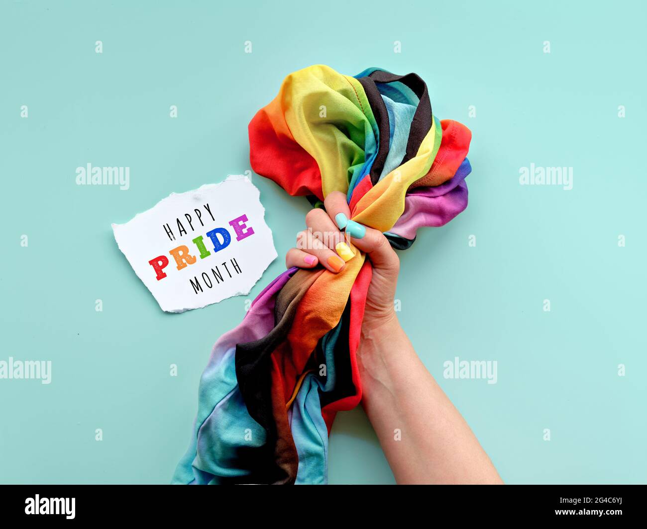LGBTQ-Community-stolz. Hand mit gefälschten Nägeln in den Farben des Regenbogens hält Regenbogen-T-Shirt. Flach liegend, Draufsicht, mintgrüner Hintergrund. Einfach, minimal Stockfoto