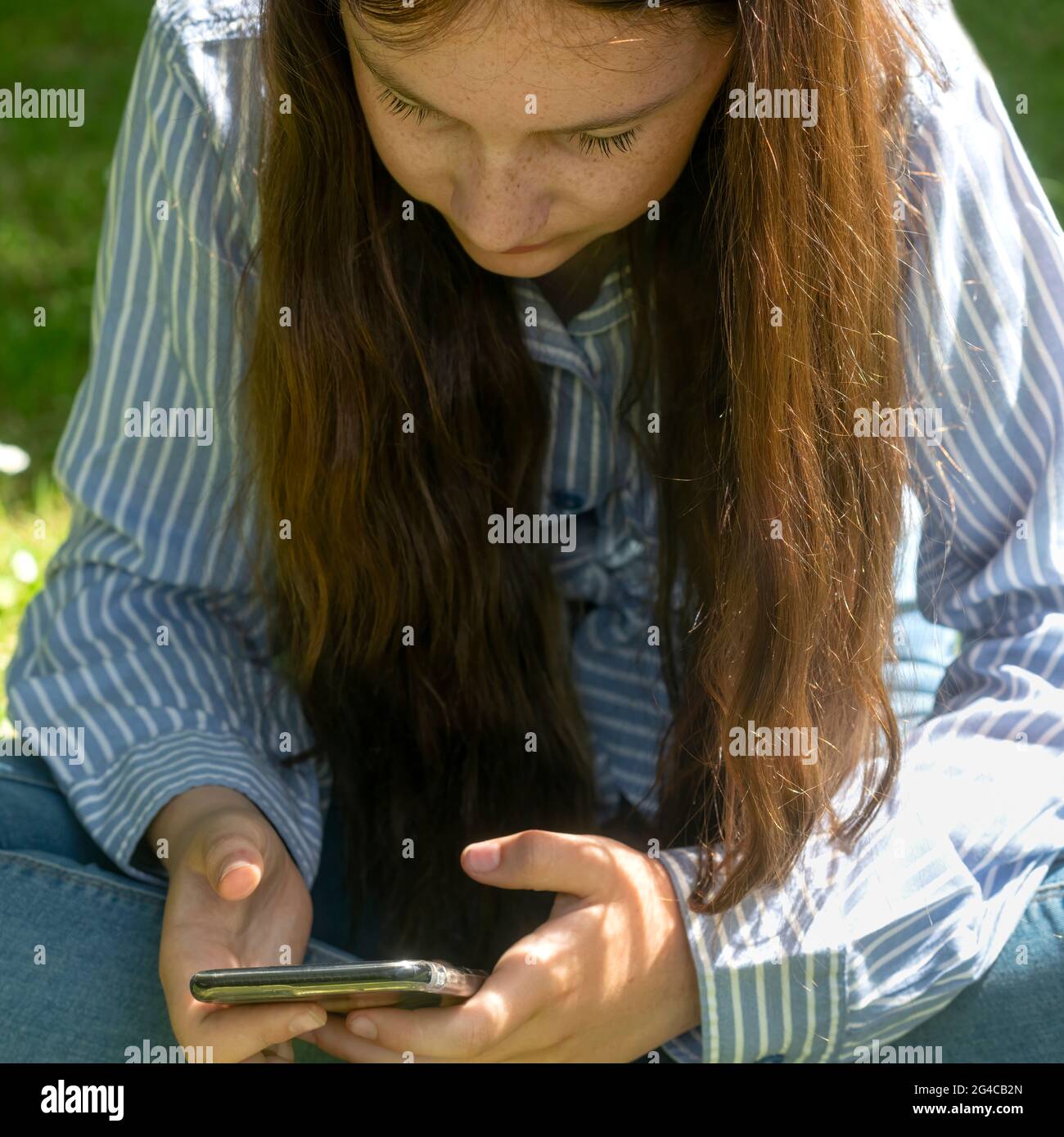 Crop weibliche Teenager mit langen Haaren sitzen auf dem Rasen und Surfen Handy an sonnigen Sommertag im Park Stockfoto