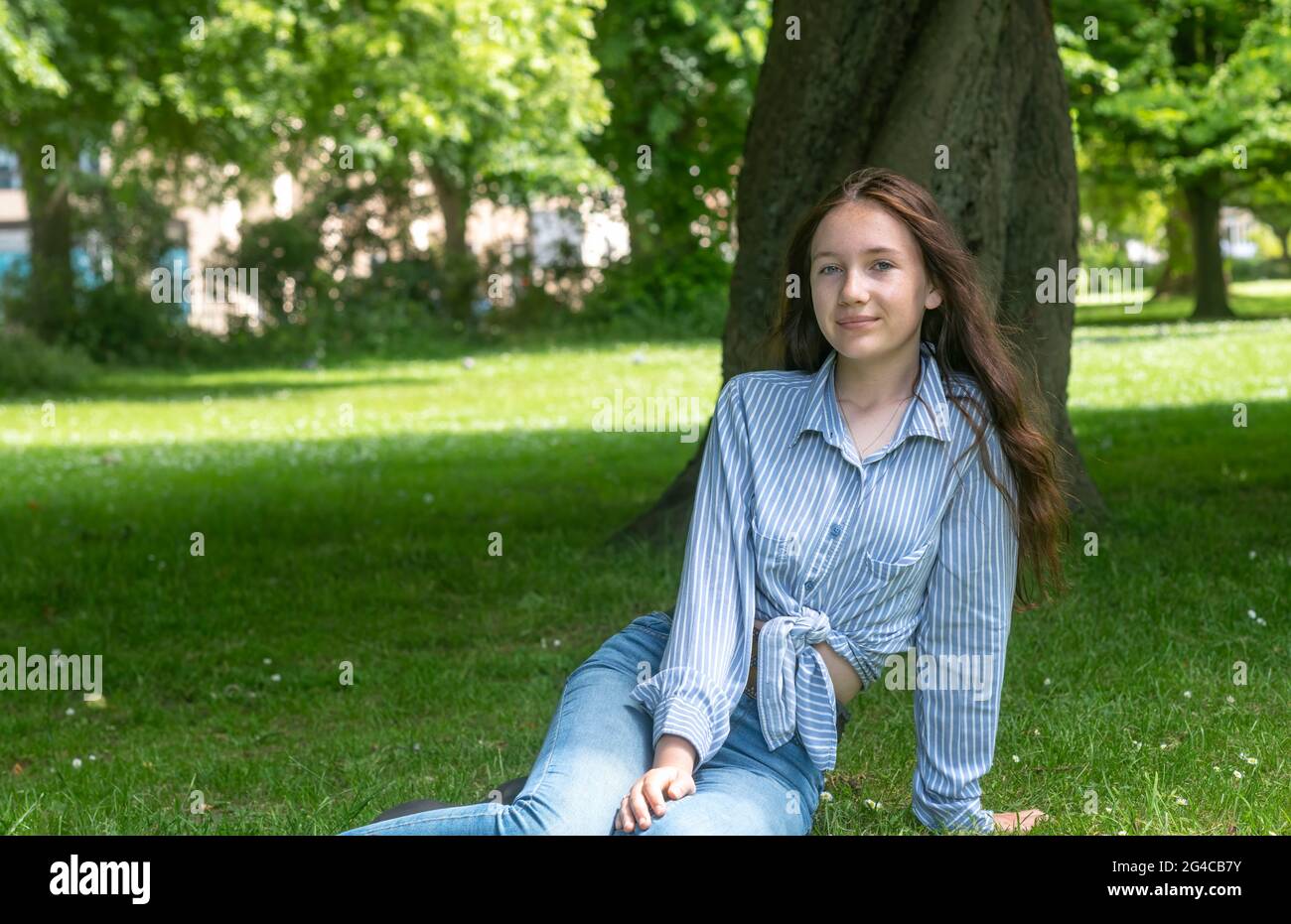 Junge Hündin im gestreiften Hemd beim Blick auf die Kamera, während sie sich im Sommer im Park auf einem Rasen in der Nähe eines Baumes ausruhte, kopieren Sie den Platz auf der linken Seite unscharfer Hintergrund Stockfoto