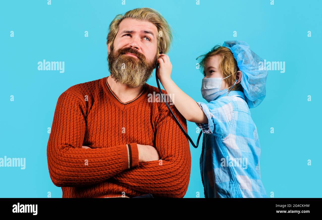 Kleiner Sohn in medizinischer Maske Spiel als Arzt mit Vater. Nettes Kind Junge vortäuschend Doktor mit Stethoskop, das Vater Patient hört. Stockfoto