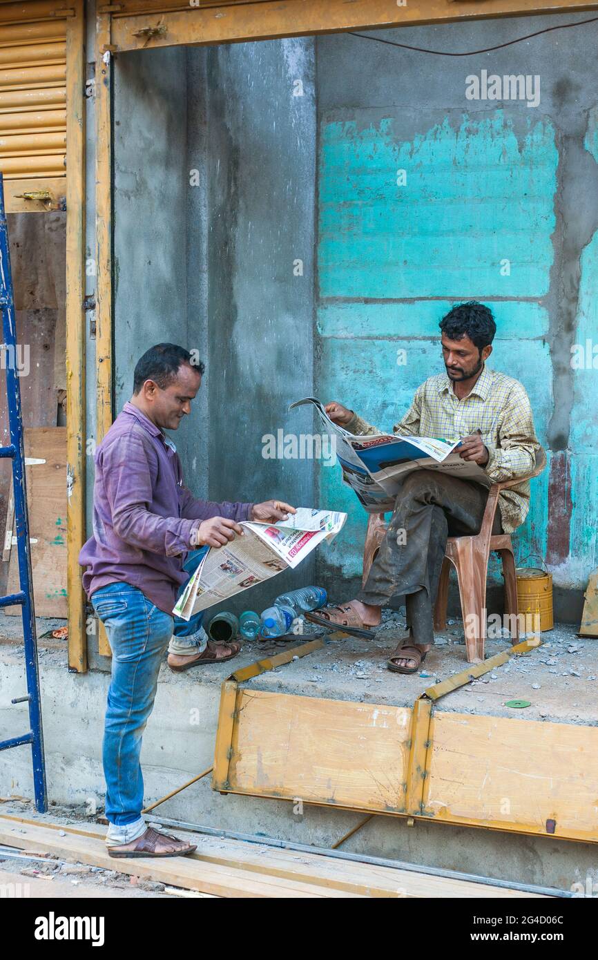 Zwei indische Arbeiter machen eine Pause und lesen die Zeitung - einer steht, der andere sitzt auf der Plattform, Gokarna, Karnataka, Indien Stockfoto