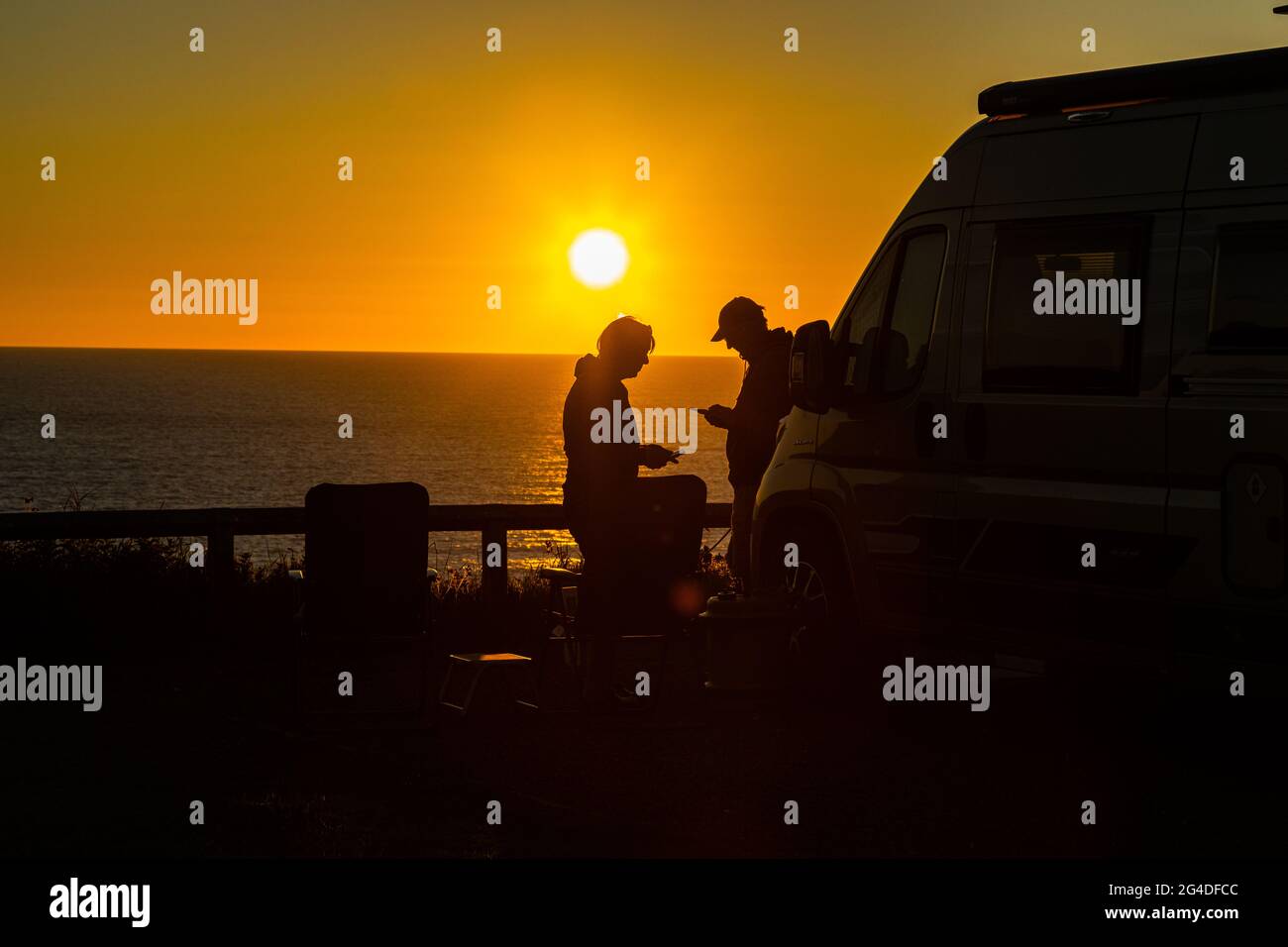 Wunderschöner Sonnenuntergang in der goldenen Stunde mit einem Paar, das sich neben einem Wohnmobil entspannt und den Sommeruntergang in Cornwall, England, genießt Stockfoto