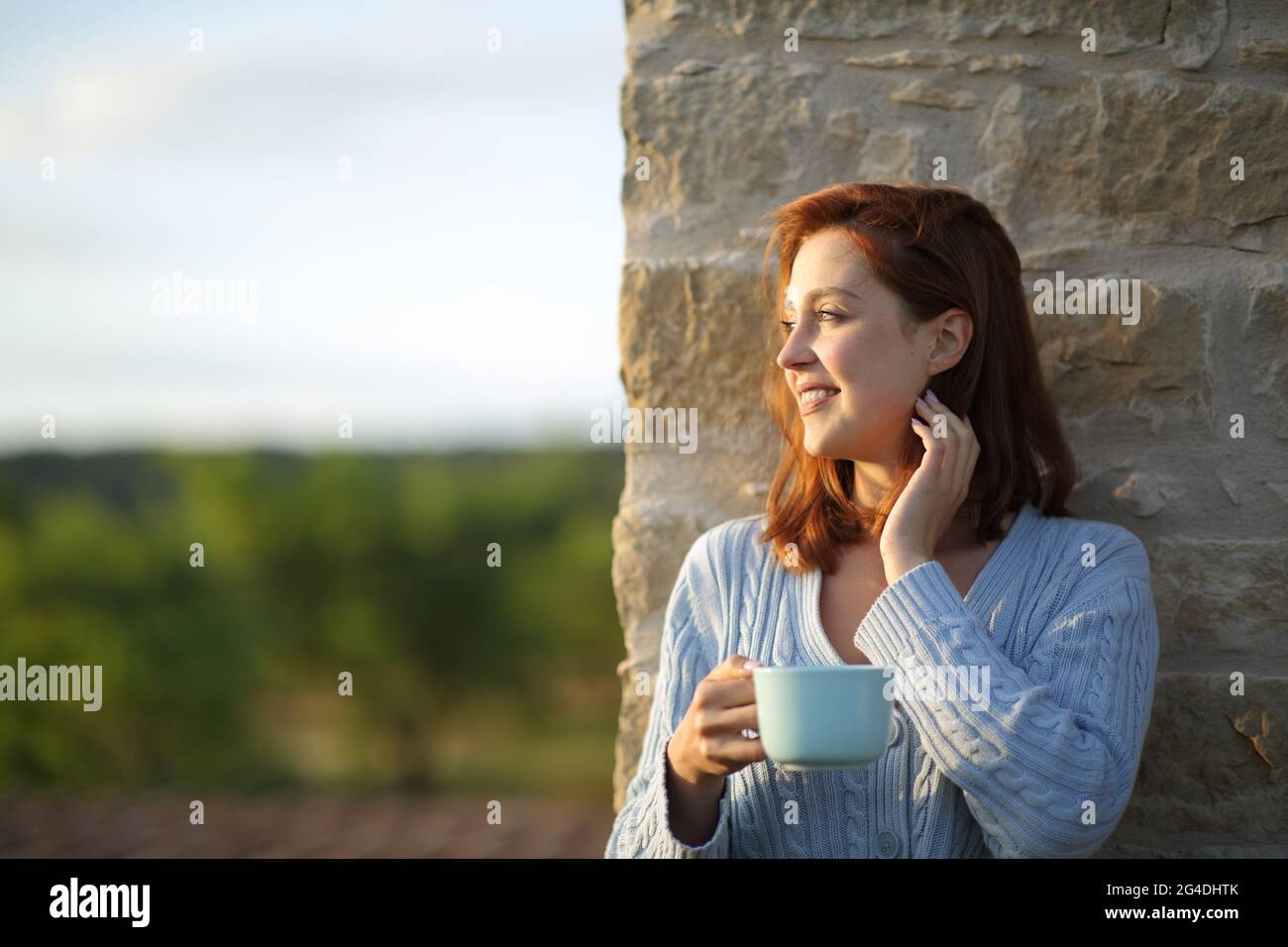 Glückliche Frau bei Sonnenuntergang, die Kaffee trinkt und die Aussicht vom Balkon aus betrachtet Stockfoto