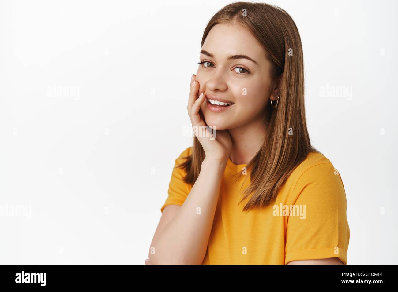 Porträt eines schönen Teenagers, Haut ohne Akne berühren, kein Make-up, Wange sanft berühren und zart lächelnd vor der Kamera, im gelben T-Shirt stehend Stockfoto