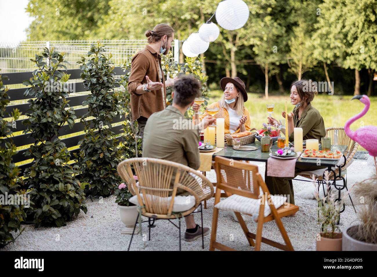 Junge Freunde in Gesichtsmaske bei einem festlichen Abendessen während eines Sommers im Hinterhof des Landhauses. Glücklich, sich im wirklichen Leben danach zu treffen Stockfoto