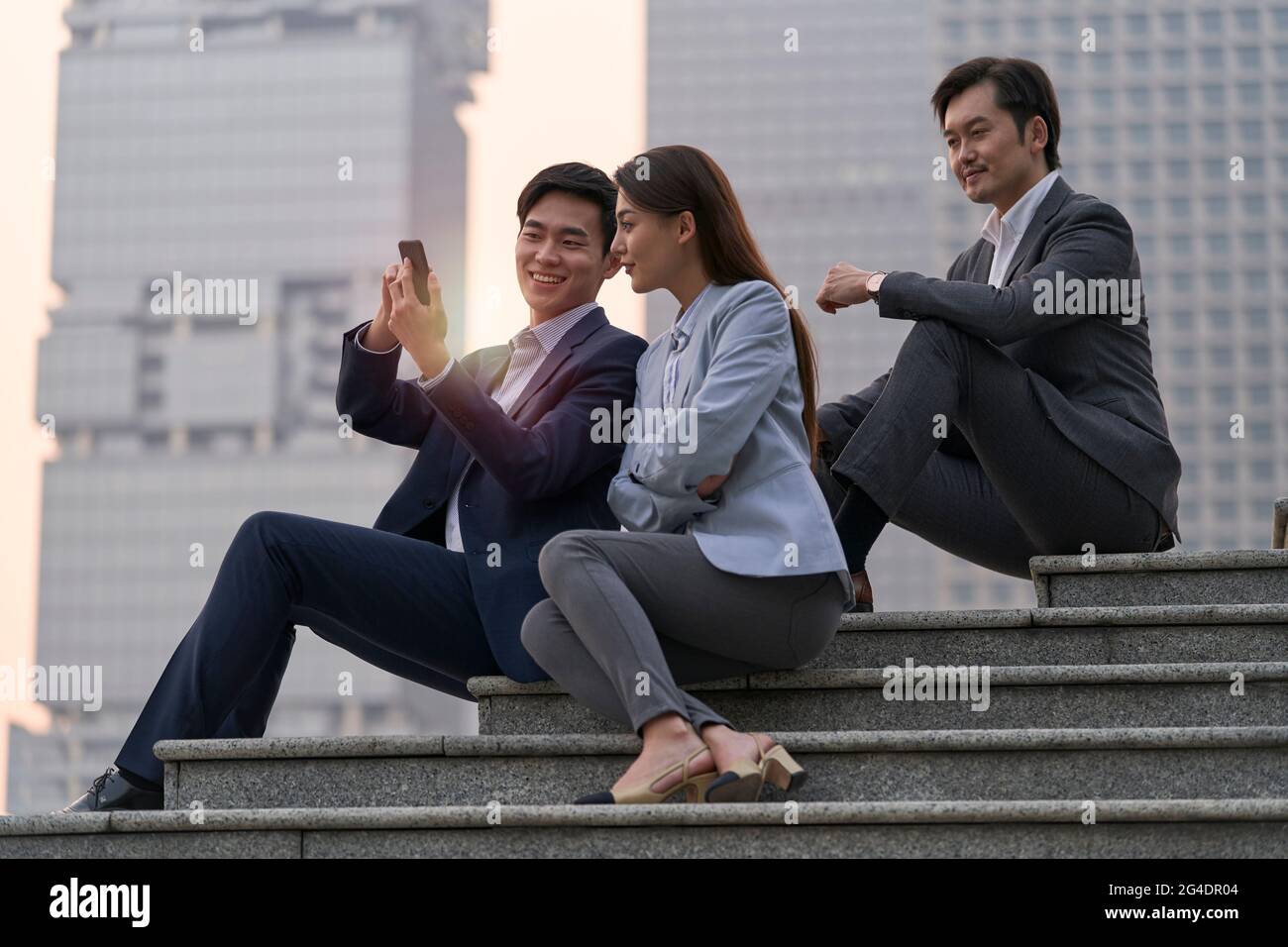 Das Team aus drei asiatischen Führungskräften sitzt auf einer Treppe und blickt auf ein Mobiltelefon mit Wolkenkratzer-Hintergrund Stockfoto