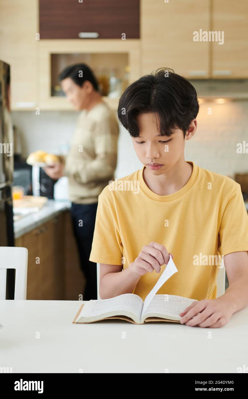 Ernsthafter Teenager, der Buch liest, als sein Vater im Hintergrund das Frühstück kocht Stockfoto